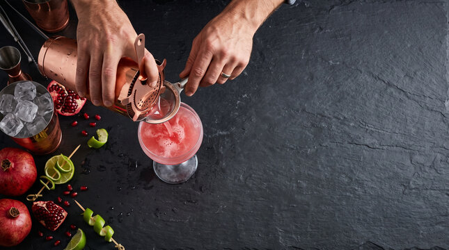 Top down view of bartender hands straining pink cocktail into glass on slate