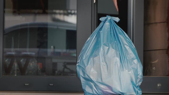 Unrecognized individual hangs a blue plastic bag of recyclables on a door handle from the outside of a house.