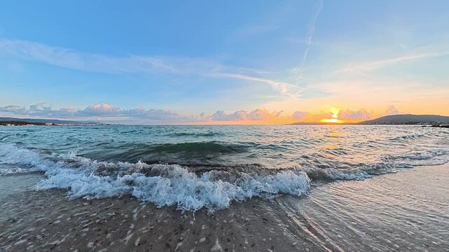 Horizontal slow motion view of small waves gently breaking along the shoreline at sunset, with warm golden light and a peaceful Mediterranean coastal atmosphere in Sardinia.