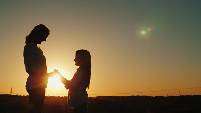 Silhouette of mother and daughter holding hands and hugging against beautiful sunset sky during golden hour.