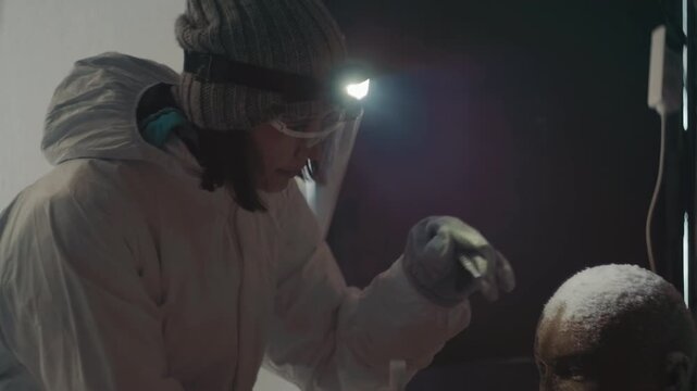 Scientist in protective suit inspects frostcovered child in dim abandoned shelter, headlamp illuminating gloved hands collecting sample with tweezers forensic researcher assessing contamination,