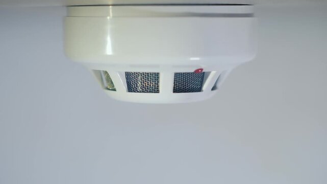 Close-up of a white smoke detector on a ceiling emitting smoke and then a red flashing light indicating alarm for fire emergency.
