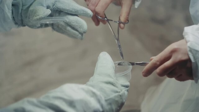 Gloved technician carefully places sample into plastic cup using clamp and tweezers, cold interior atmosphere with frost and dust, closeup of coordinated hands performing precise exchange