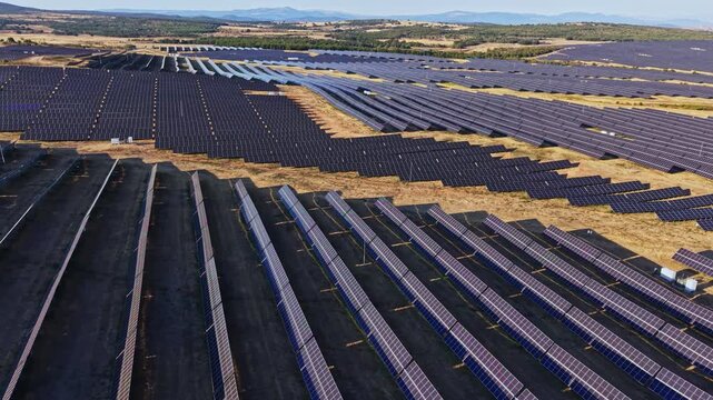 A large solar panel farm is located in a rural area with rows of solar panels aligned in different directions. The sun shines on the panels as they generate energy.