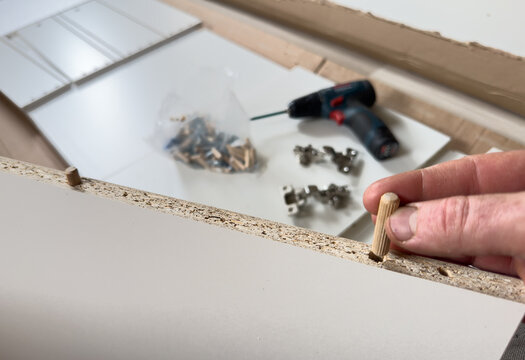 Hand inserting wooden dowel into particleboard furniture panel during assembly, with drill and hardware visible in background