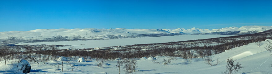 Enontekiö, Finland - 02.04.2023: Panoramic view of snow-covered mountains and frozen lake under a clear bright blue arctic sky © Andrei