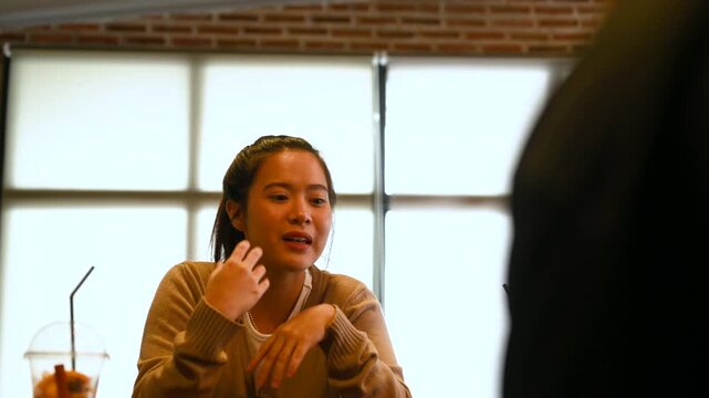 A woman gestures in conversation with another person across a cafe table. The woman faces the other diner as a laptop, cold drinks, glassware, and brick walls surround the wooden table.
