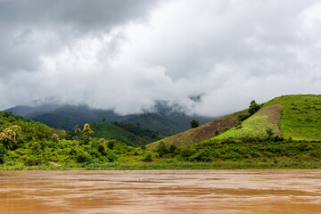Fototapeta premium Muddy brown Mekong river water flows past dense tropical vegetation in Laos under a sunny sky.