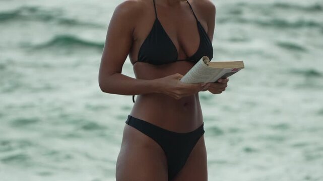 Beach reader focused on paper book near surf, enjoying tropical vacation sunlight. Static medium shot captures body movement against ocean.