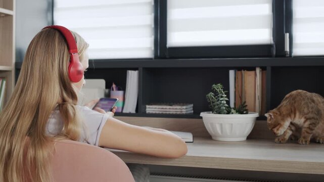 Young blonde woman in red headphones sits at table and uses smartphone, curious cat approaches flower pot on desk, domestic animals in home interior.