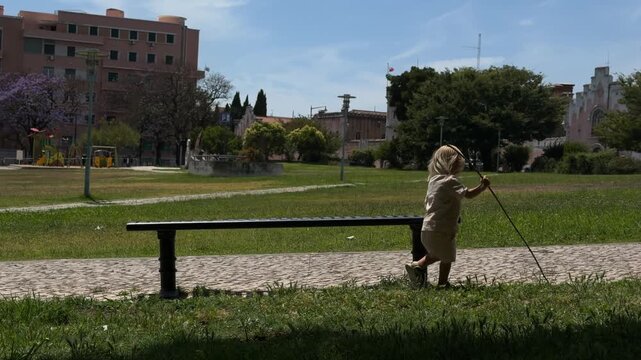 Young child walking past metal park bench on cobblestone path, holding long stick overhead, urban park playground and trees under bright midday blue sky