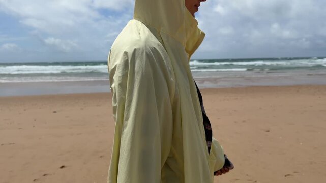 Person in pale yellow hooded jacket walking along cloudy sandy shoreline, raising hood and adjusting jacket collar as rolling surf breaks in background