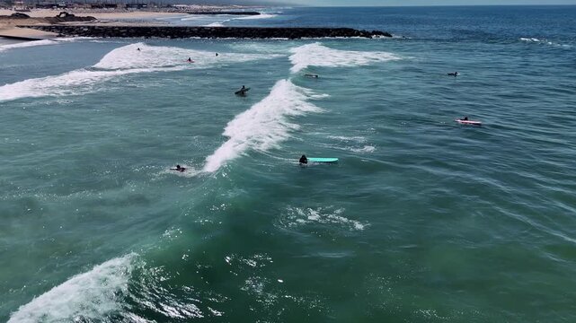 Coastal surfers on paddleboards waiting in turquoise water beside rocky breakwater near shoreline under bright midday sun with foam crests and sun glints along ocean horizon