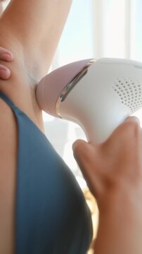 Handheld hair removal device pressing against underarm skin during at home grooming demonstration, close angled view with warm natural window backlight and soft highlights