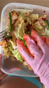 Gloved hand mixing seasoned napa cabbage and green onion for kimchi in clear plastic container with chili paste smears, close overhead view on light wood tabletop