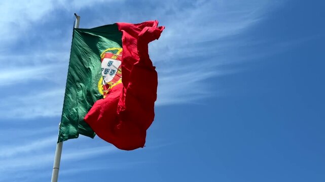 Low-angle view of national flag fluttering in strong wind, red and green fabric with central yellow emblem and partial white shield, waving across bright blue sky with soft clouds