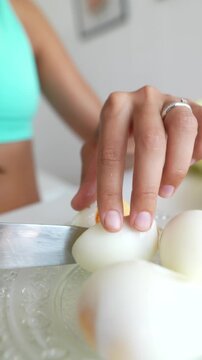Close-up hand slicing peeled hard-boiled egg on decorative glass plate with large kitchen knife, showing cut line and yolk fragments beside more peeled eggs