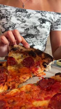 Hands lifting pepperoni pizza slice with charred crust, gooey cheese stretch, glossy tomato sauce, and blistered browned bite, fingers in floral off-shoulder top under warm indoor lighting