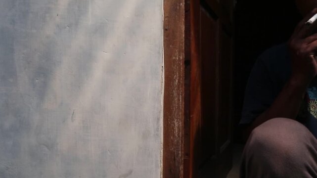 A Javanese man sits casually in front of his house, smoking a kretek clove cigarette during a quiet moment at the doorway.
