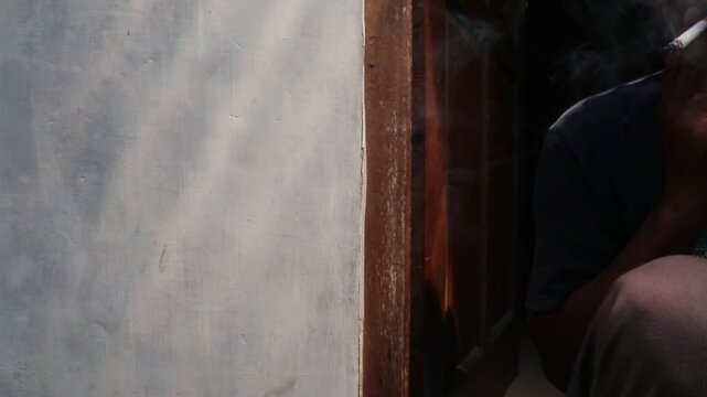 A Javanese man sits casually in front of his house, smoking a kretek clove cigarette during a quiet moment at the doorway.