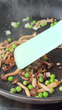 Close-up frying pan stir of browned mushrooms, green peas, and diced onion with spatula on stovetop