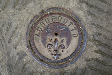 Rusty iron manhole cover with Giglio fiorentino fleur de lis crest and word ACQUEDOTTO or Aqueduct, marking the city water system in Florence, Italy © SvetlanaSF