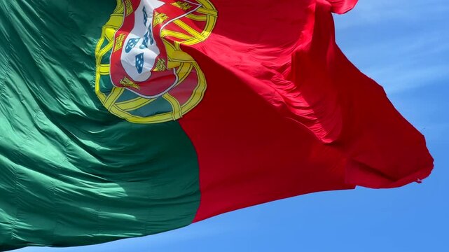 Low-angle view of national flag fluttering in strong wind, red and green fabric with central yellow emblem and partial white shield, waving across bright blue sky with soft clouds