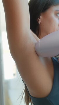 Close-up of woman underarm grooming with handheld hair removal device against raised armpit, warm smooth skin and light stubble shown in soft window daylight indoors