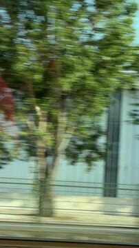 Motion blurred train travel perspective with steel rails and concrete barrier in foreground, trimmed hedge and street lamp midground, red brick station building with arched windows under blue sky