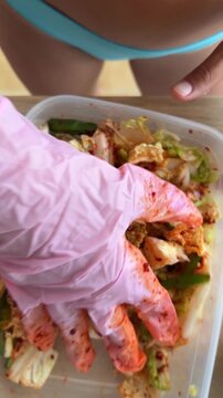 Pink gloved hand mixing and packing napa cabbage kimchi into rectangular plastic container on wooden table with turquoise garment background overhead