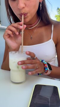 Young woman stirring and tasting iced milk drink at outdoor cafe table, paper straw in glass with ice and condensation, layered necklaces, pink choker, rings and bracelets