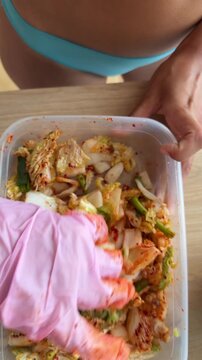 Pink disposable gloved hand mixing spicy napa cabbage in clear plastic container on wooden table, with turquoise swimwear torso partially visible in background