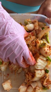 Pink gloved hand pressing and folding seasoned napa cabbage with chili paste in clear plastic container on kitchen cutting board