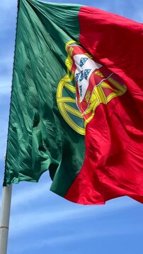 Low-angle view of national flag fluttering in strong wind, red and green fabric with central yellow emblem and partial white shield, waving across bright blue sky with soft clouds