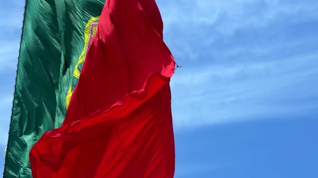 Low-angle view of national flag fluttering in strong wind, red and green fabric with central yellow emblem and partial white shield, waving across bright blue sky with soft clouds