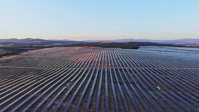 Solar panels are arranged in straight rows in a large field. The scene shows a rural area in Spain at sunset. The horizon features hills and mountains in the background.
