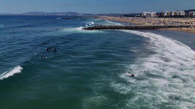 Aerial view of surfers waiting near breaking waves on crowded sandy beach under bright midday sun with umbrellas and low-rise buildings along coastline