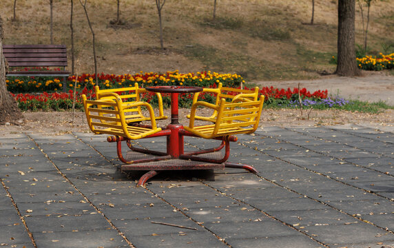 Vintage red and yellow metal carousel standing on a paved playground area in a sunny autumn park with colorful flower beds