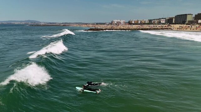 Two surfers in black wetsuits paddle surfboards through breaking waves beside rocky jetty, with crowded sandy beach and low coastal buildings under bright midday sky
