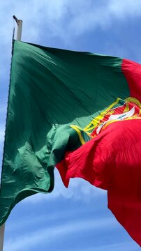 Low-angle view of national flag fluttering in strong wind, red and green fabric with central yellow emblem and partial white shield, waving across bright blue sky with soft clouds