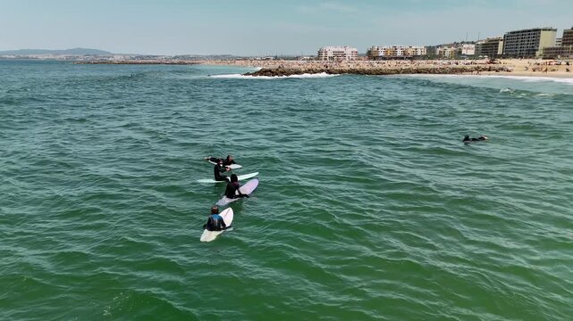 Group of surfers in dark wetsuits sitting and paddling on surfboards in green ocean near sandy beach, rocky jetty and distant beachfront buildings under bright midday sun