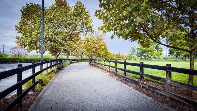 a paved pathway lined by dark wooden fences curves through a lush green park with trees under an overcast sky