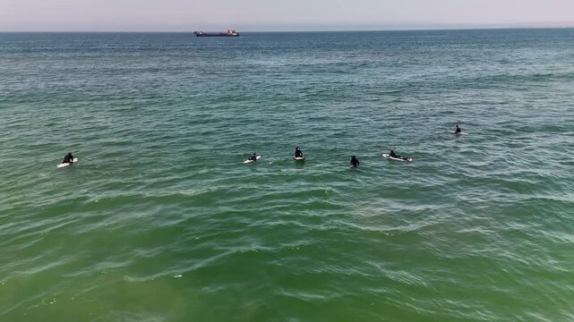 Overhead view of six surfers resting on surfboards in calm green sea, distant cargo ship on horizon under hazy sky for peaceful coastal leisure