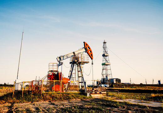 Oil pump jack and drilling rig in open field under clear blue sky. Oil extraction from a well. Oil field, oil drilling operations at rig.