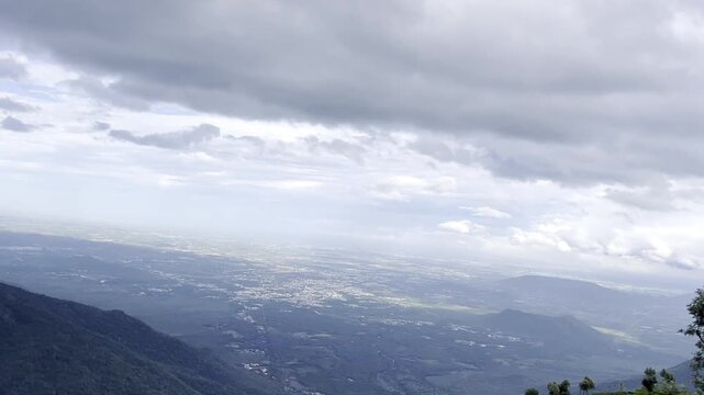 Panoramic mountain valley view from green highlands with scattered trees, tea plantation slopes and distant plains beneath a dramatic cloudy sky, creating a peaceful scenic travel landscape in nature.