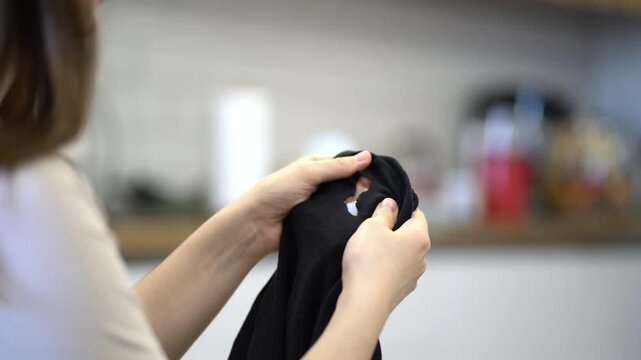 Young woman examines a hole in her black shirt indoors, gripping the damaged garment and reacting with disappointment.