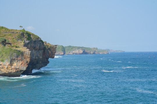 Scenic tropical coastal view with rugged rock cliffs covered in vegetation meeting vibrant blue ocean waves under a clear sky in Indonesia.
