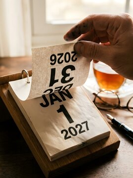 Person flipping the page of a wooden desk calendar from december 31 2026 to january 1 2027 to mark the start of a new year during a bright sunny morning in a home office workspace