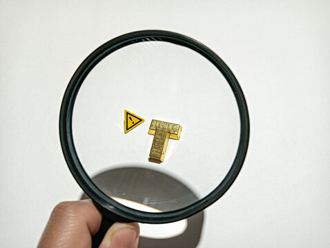 Magnifying glass focusing on a small gold T-shaped object beside a warning symbol, representing inspection, investigation, risk assessment, security, and careful examination.
