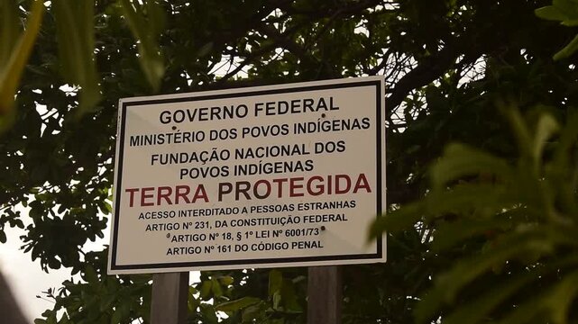 Placa de demarca&ccedil;&atilde;o de terra ind&iacute;gena em meios as folhas verdes balan&ccedil;ando, aviso de terra protegida dos povos origin&aacute;rios do brasil